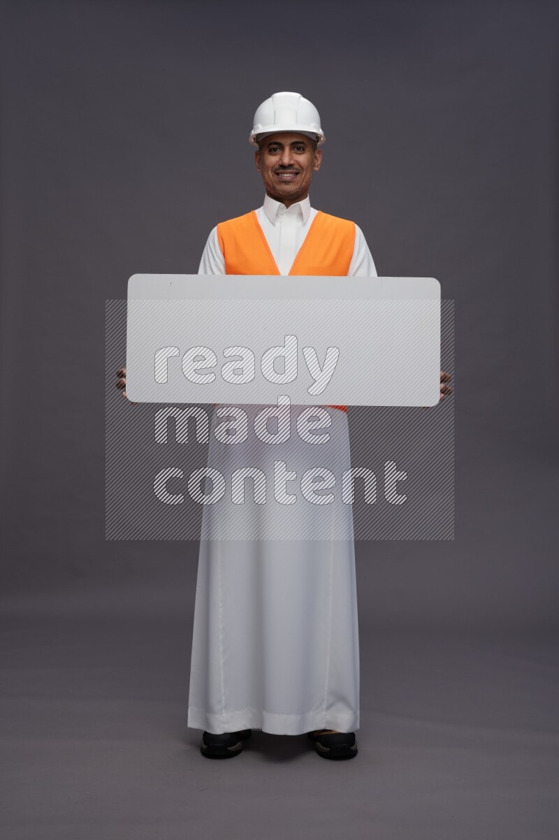 Saudi man wearing thob with engineer vest standing holding board on gray background