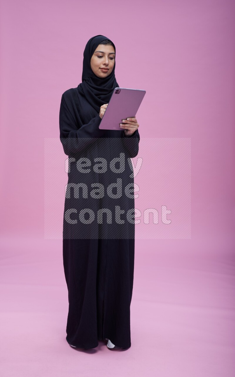 Saudi woman wearing Abaya standing working on tablet on pink background