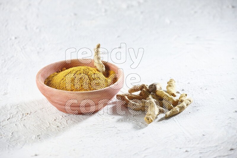 A wooden bowl full of turmeric powder and dried turmeric whole fingers beside it on textured white flooring