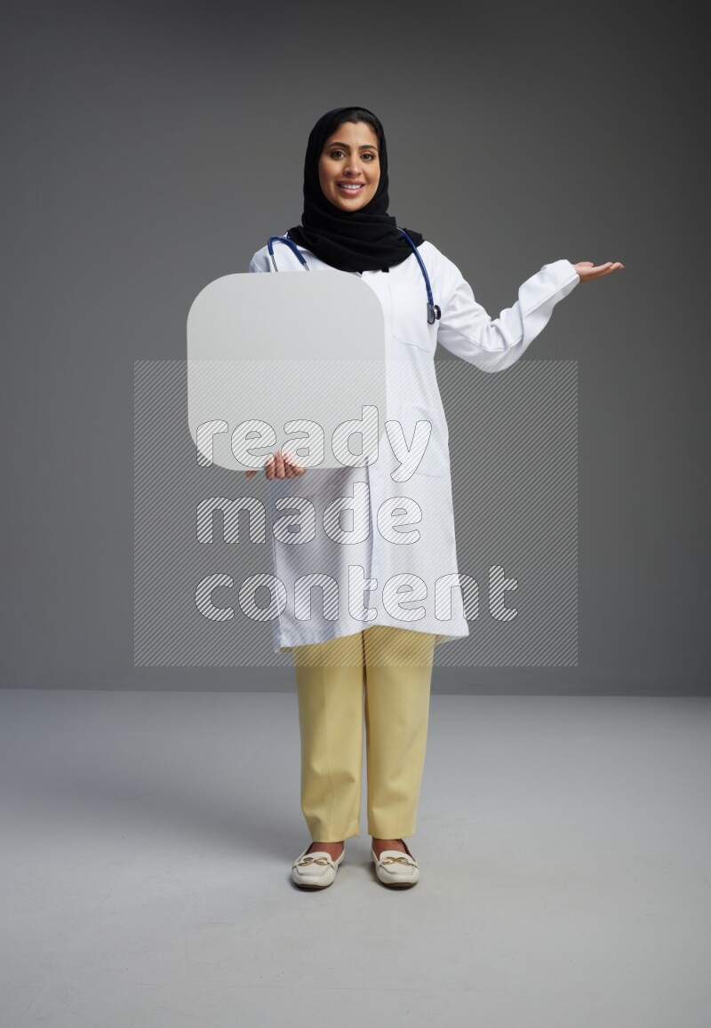 Saudi woman wearing lab coat with stethoscope standing holding social media sign on Gray background