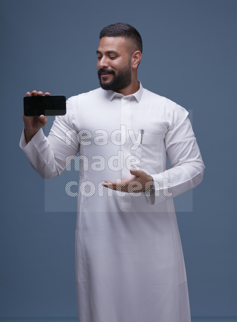 A man Showing his phone in Blue Background wearing Saudi Thob