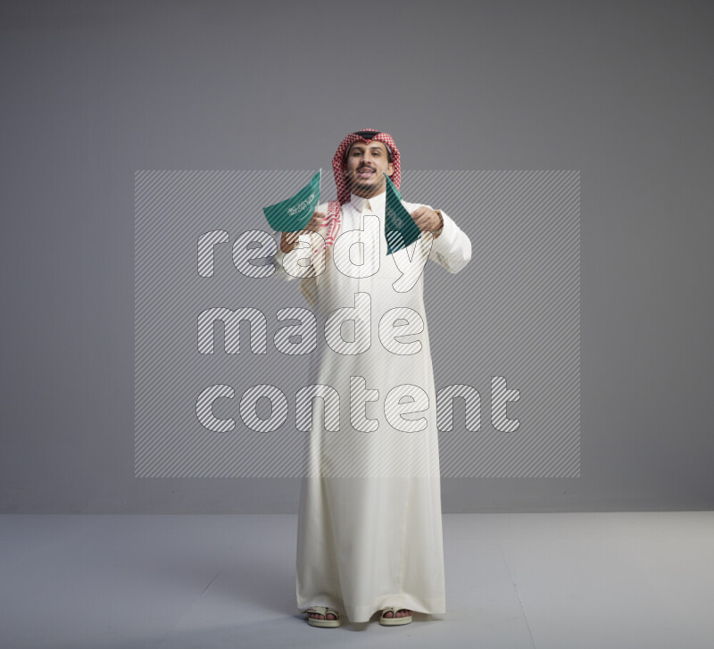 A Saudi man standing wearing thob and red shomag raising small Saudi flag on gray background