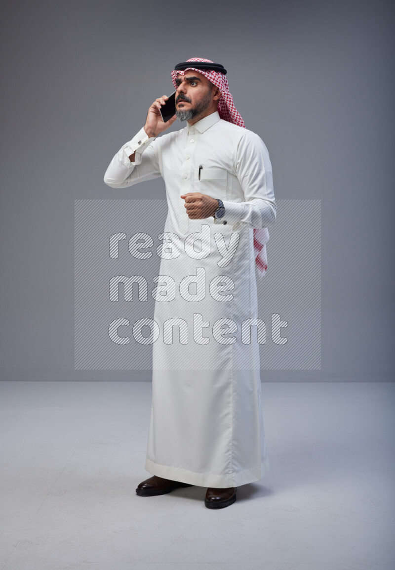 Saudi man Wearing Thob and red Shomag standing talking on phone on Gray background