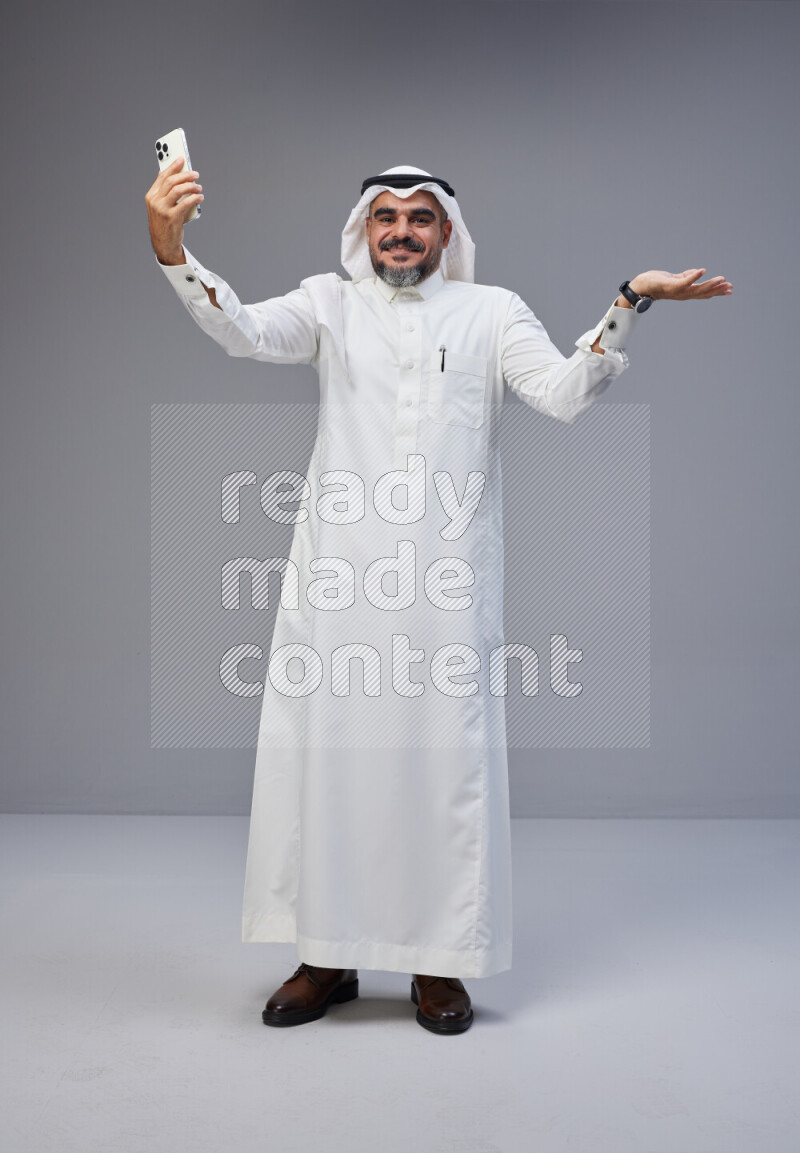 Saudi man Wearing Thob and white Shomag standing taking selfie on Gray background