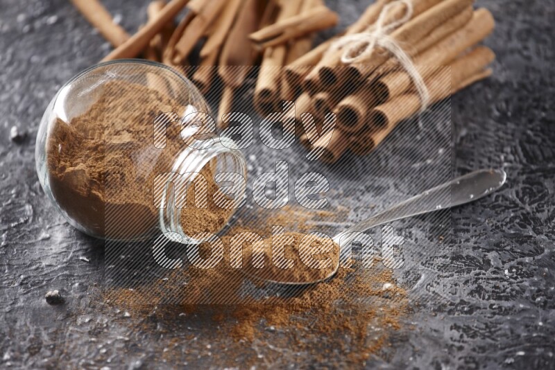 Herbal glass jar full cinnamon powder flipped and a metal spoon full of powder, cinnamon sticks stacked and bounded in the back on textured black background in different angles