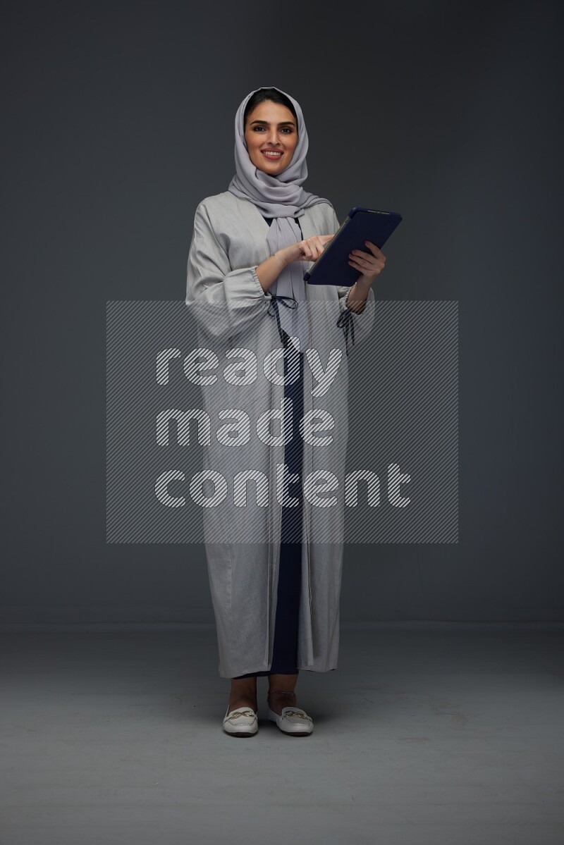 A Saudi woman wearing a light gray Abaya and head scarf standing holding a phone and pointing with the other hand eye level on a grey background