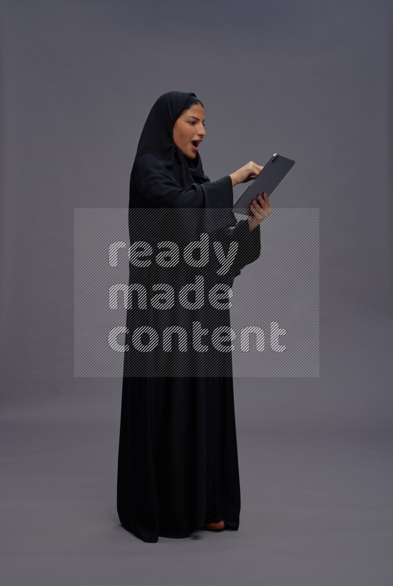 Saudi woman wearing Abaya standing working on tablet on gray background