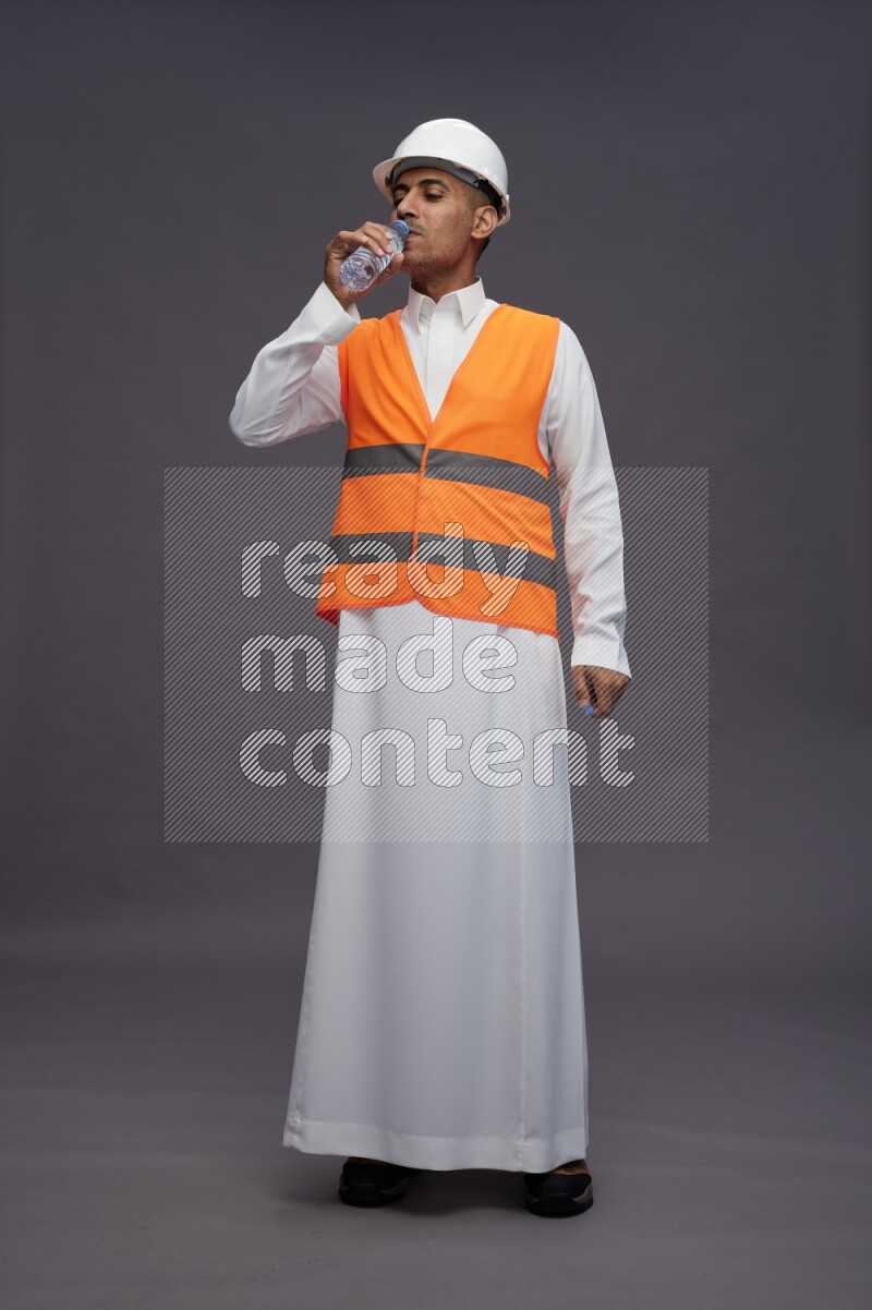 Saudi man wearing thob with engineer vest standing drinking water on gray background