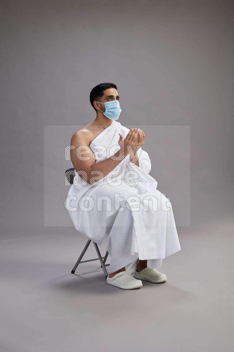 A man wearing Ehram with face mask sitting on chair dua'a on gray background