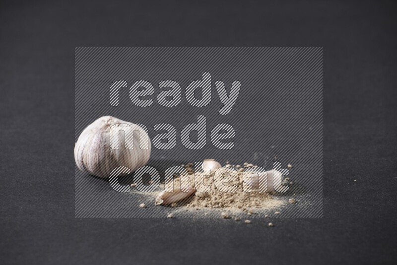 A small pile of garlic powder with some of cloves and a whole garlic bulb on a black flooring