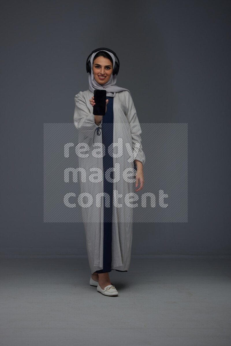 A Saudi woman wearing a light gray Abaya and head scarf setting on a dark grey chair and watching on the tablet eye level on a grey background