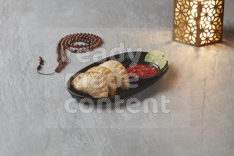 Three fried sambosas in an oval shaped black plate and a red sauce in a black round ramekin with a brown misbaha and a golden lantern on the side on a gray background