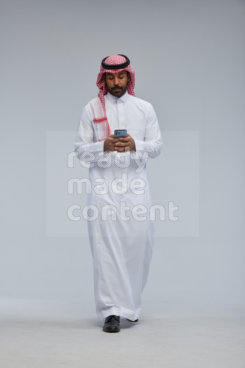 Saudi man Wearing Thob and shomag standing texting on phone on Gray background