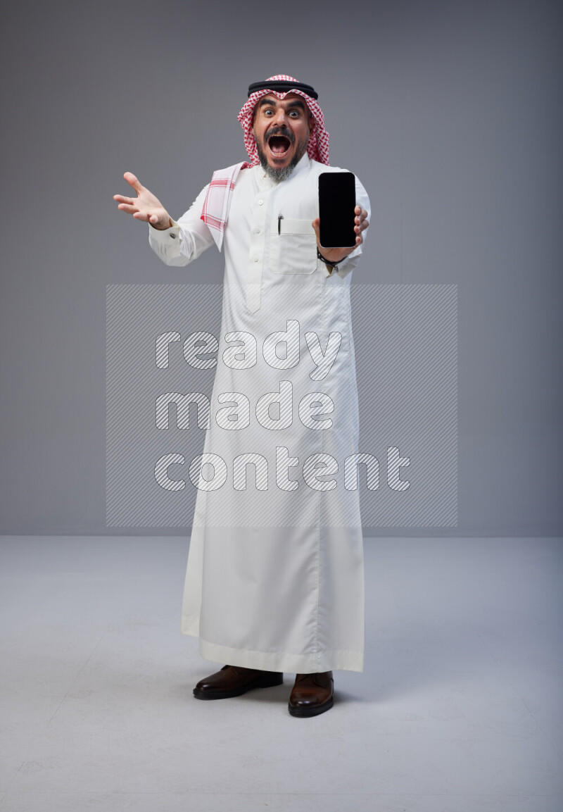 Saudi man Wearing Thob and red Shomag standing showing phone to camera on Gray background