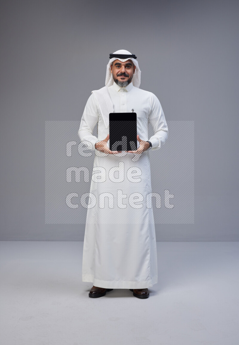 Saudi man Wearing Thob and white Shomag standing showing tablet to camera on Gray background
