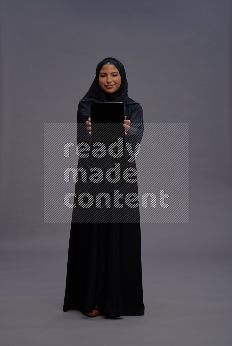 Saudi woman wearing Abaya standing showing tablet to camera on gray background