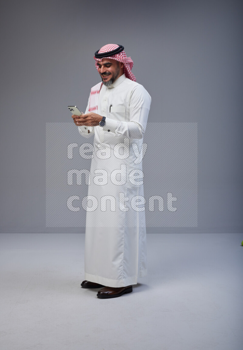 Saudi man Wearing Thob and red Shomag standing texting on phone on Gray background