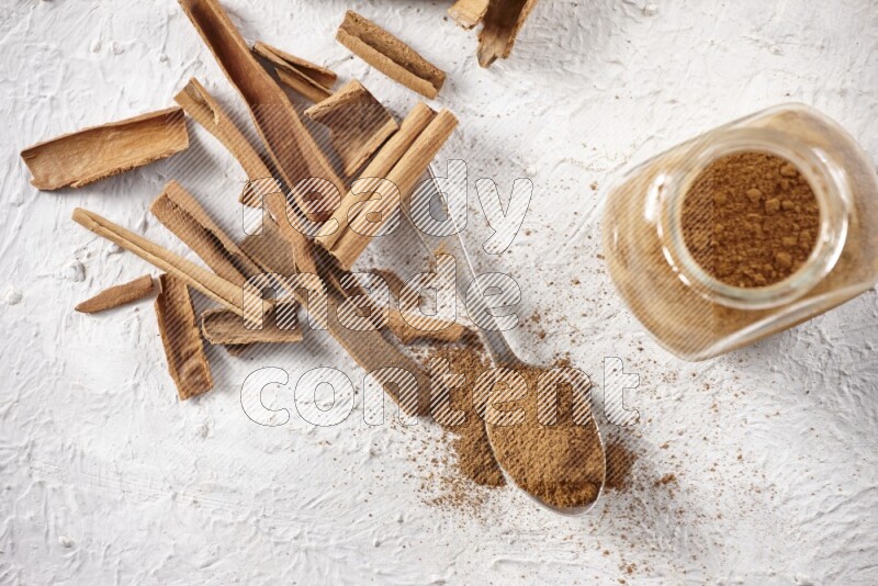 Herbal glass jar full cinnamon powder and a metal spoon surrounded by cinnamon sticks on a white background