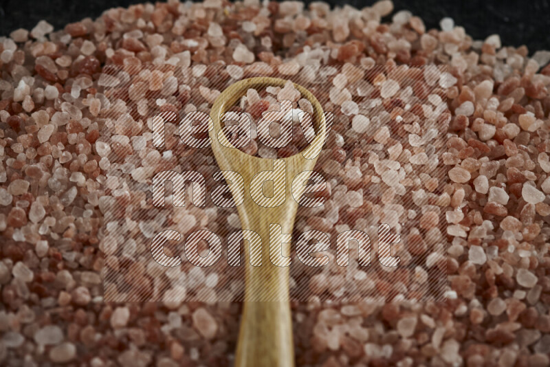 A wooden spoon full of coarse himalayan salt crystals on a bunch of the crystals on black background