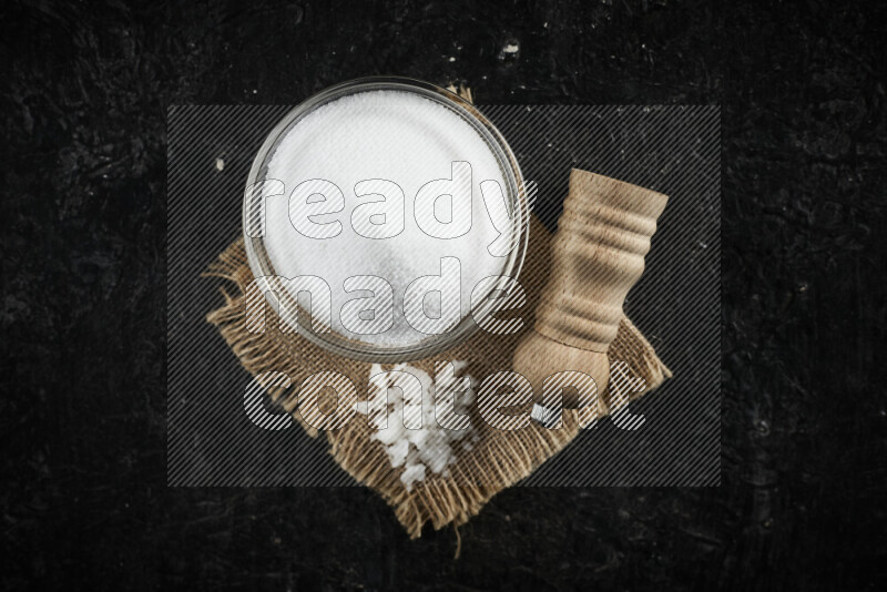 A glass bowl full of white salt with a wooden grinder on a burlap fabric all on black background