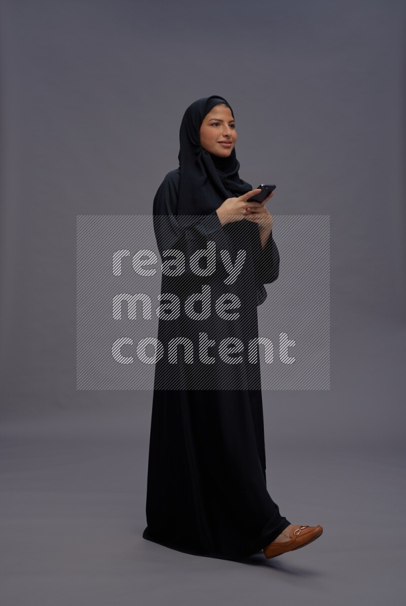 Saudi woman wearing Abaya standing texting on phone on gray background