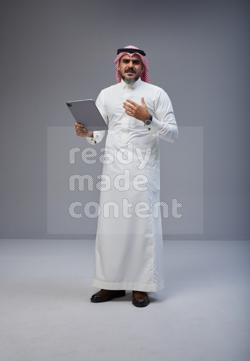 Saudi man Wearing Thob and red Shomag standing working on tablet on Gray background