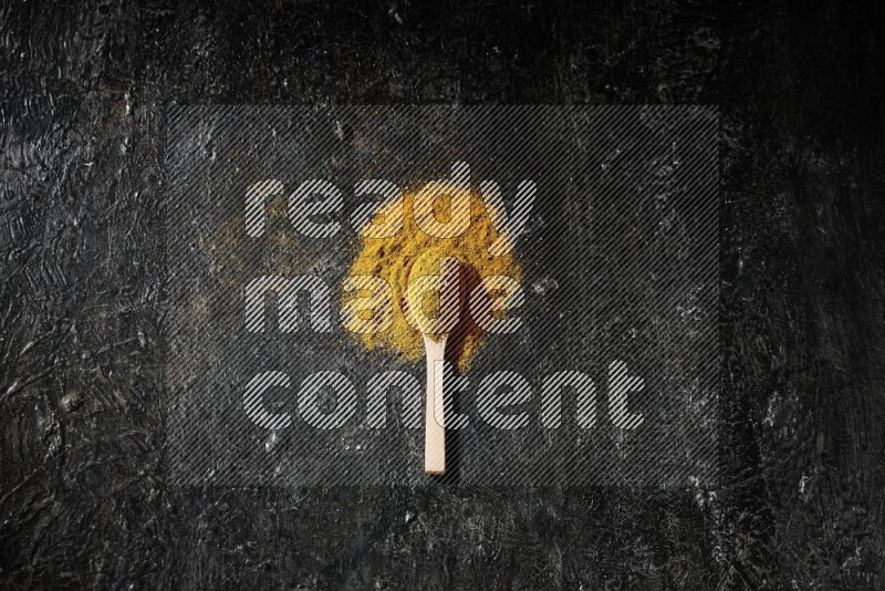 A wooden spoon full of turmeric powder on textured black background