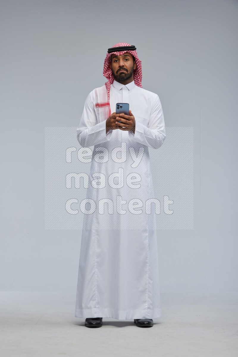 Saudi man Wearing Thob and shomag standing texting on phone on Gray background