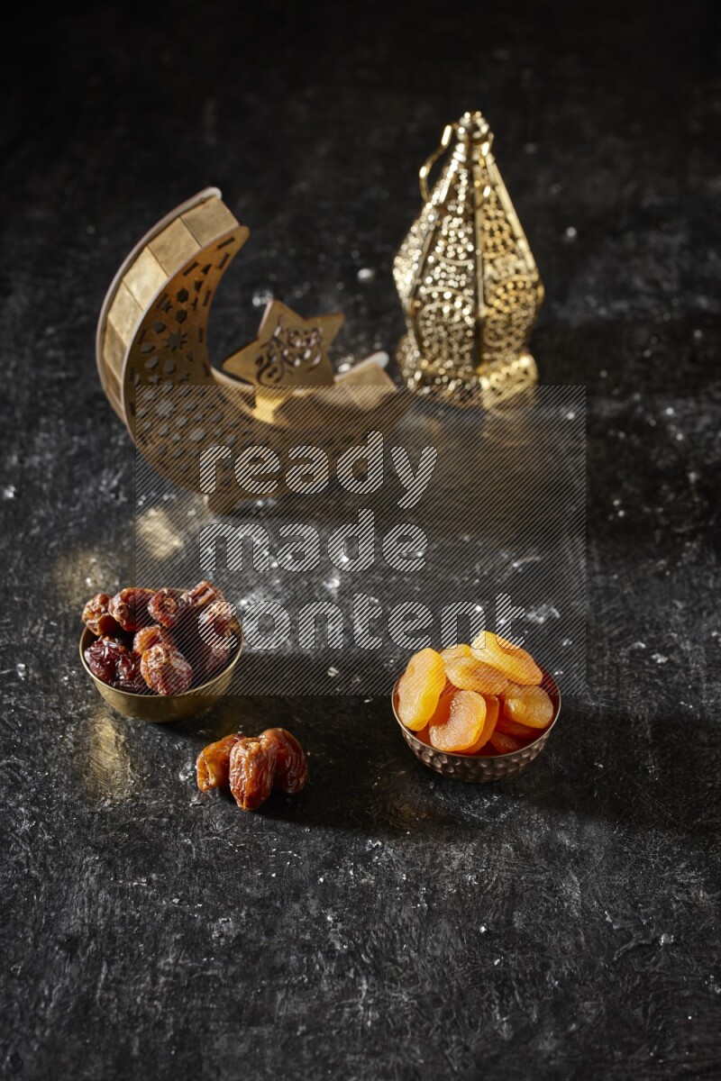 Dates in a metal bowl with dried apricot beside golden lanterns in a dark setup