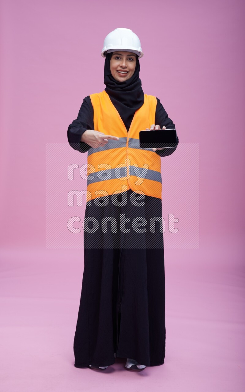 Saudi woman wearing Abaya with engineer vest and helmet standing showing phone to camera on pink background
