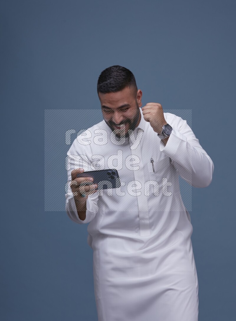 A man Playing a Game on his Smartphone  on Blue Background wearing Saudi Thob