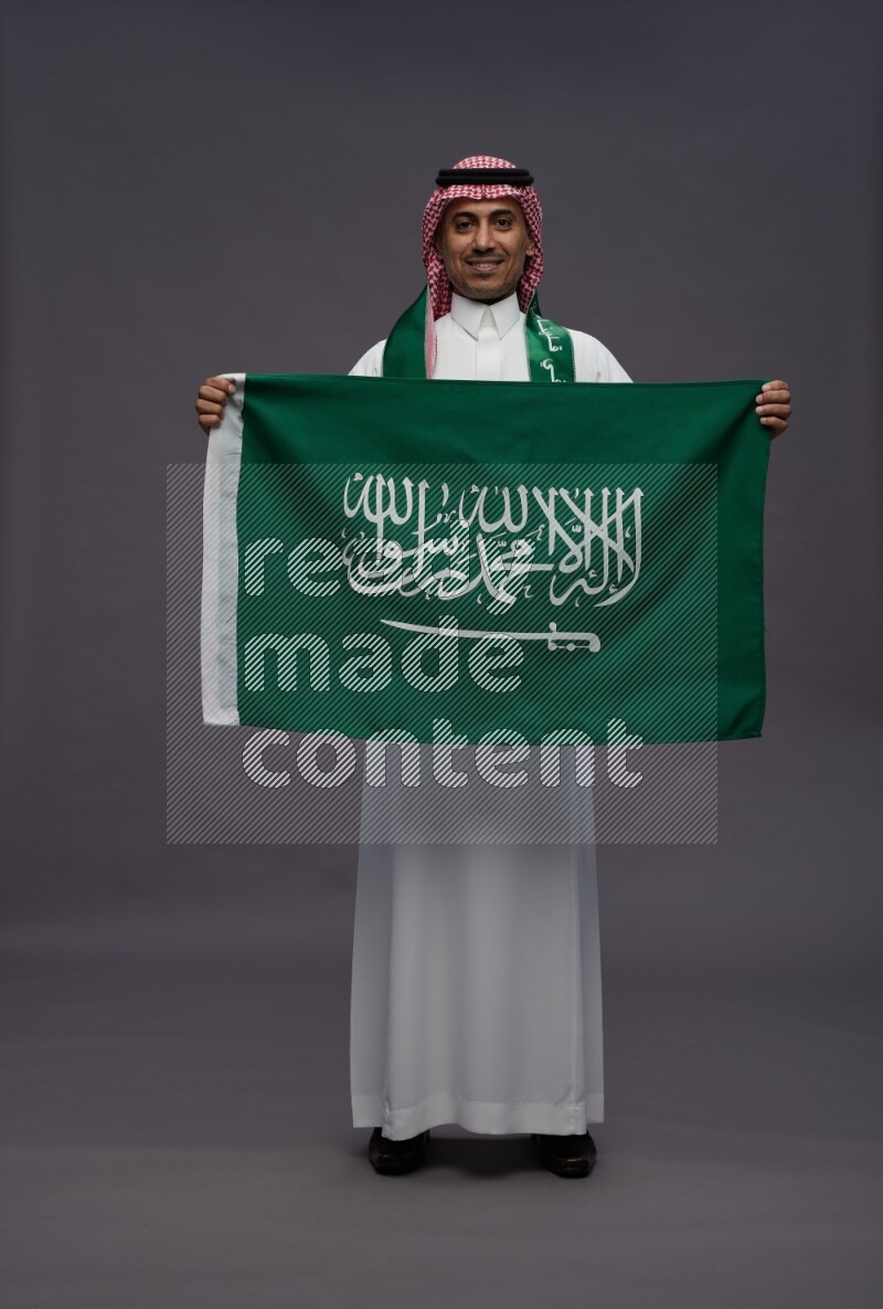 Saudi man wearing thob and shomag standing holding Saudi flag on gray background