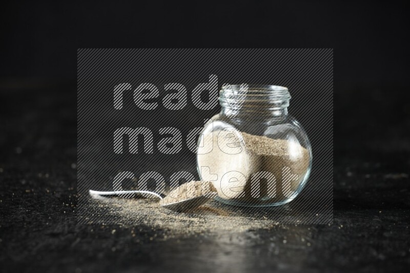 A glass spice jar and metal spoon full of cardamom powder on textured black flooring