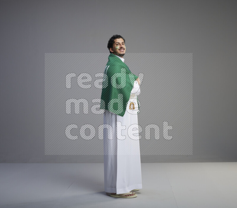 A Saudi man standing wearing thob and saudi flag scarf with face painting wrapping big Saudi flag on gray background