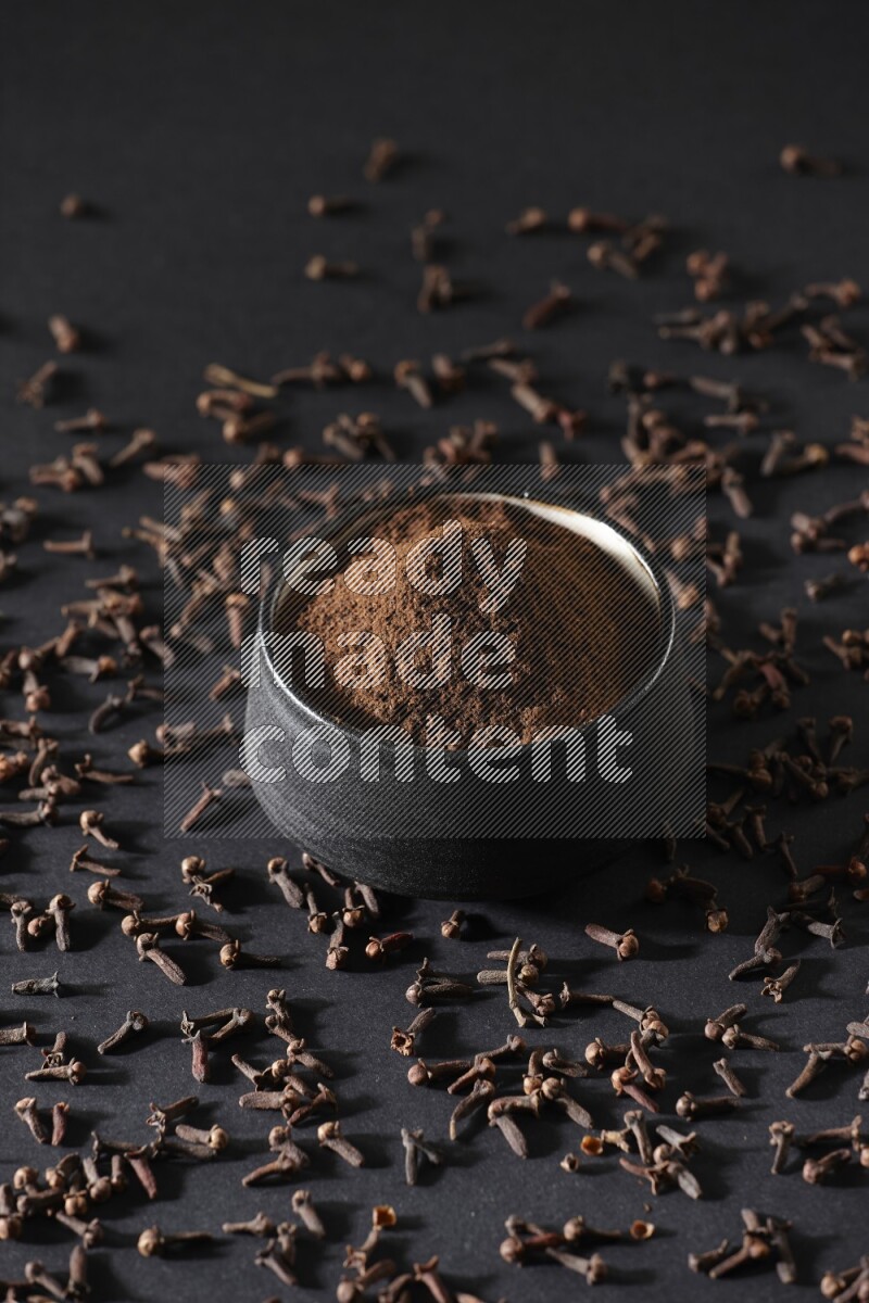A black pottery bowl full of cloves powder and cloves whole grains on a black flooring