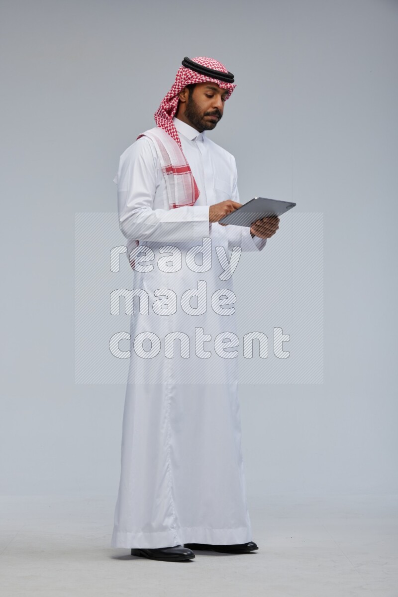 Saudi man Wearing Thob and shomag standing working on tablet on Gray background