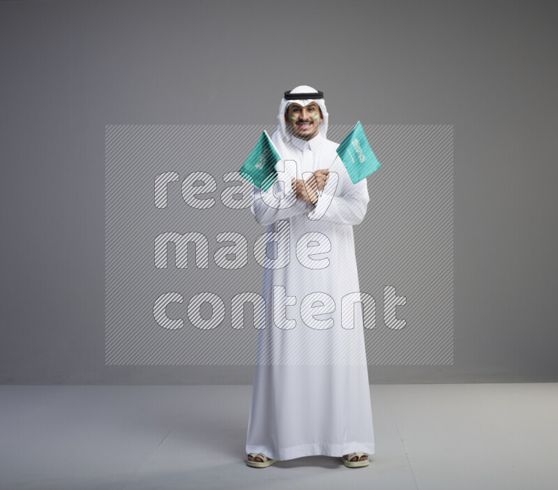 A Saudi man standing wearing thob and white shomag with face painting raising small saudi flag on gray background