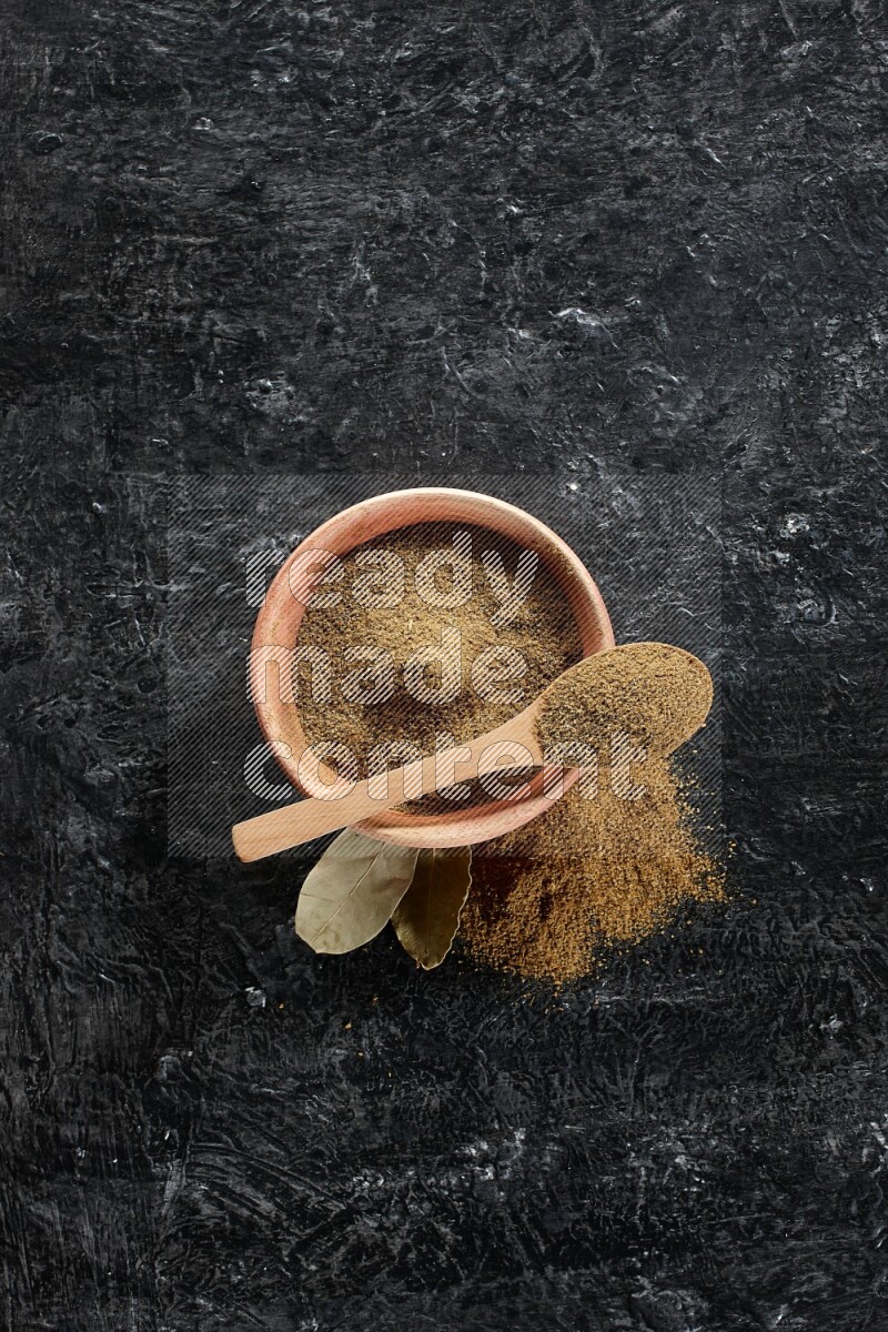 A wooden bowl and spoon full of cumin powder on a textured black flooring