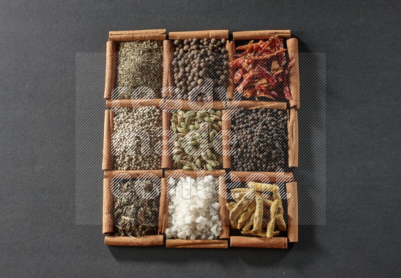9 مربعات من أعواد القرفة ممتلئة بالهيل في المنتصف محاطة بأصابع الكركم والفلفل الأسود والفلفل الحار والبهار الحلو والكمون والفلفل الأبيض والريحان المجفف والملح على أرضية سوداء