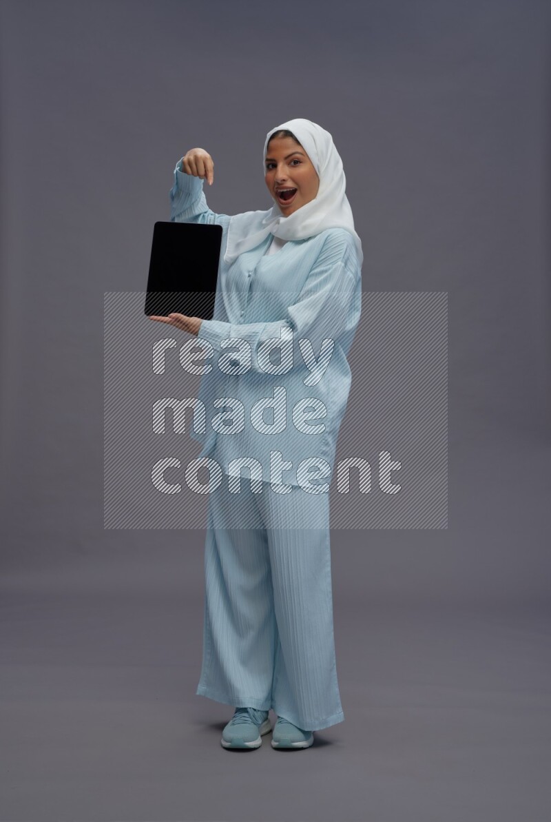 Saudi woman wearing hijab clothes standing showing tablet to camera on gray background
