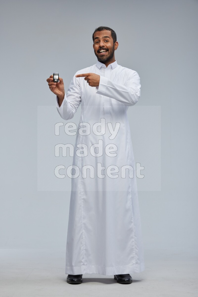 Saudi man wearing thob and shomag standing holding car key on gray background