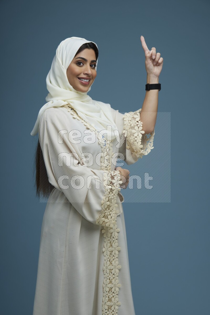 A Saudi woman pointing in a blue background wearing an off-white Abaya Hijab
