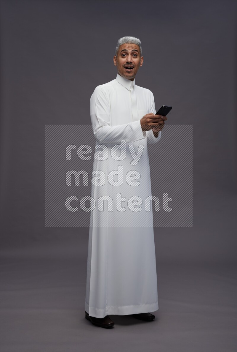 Saudi man wearing thob standing texting on phone on gray background