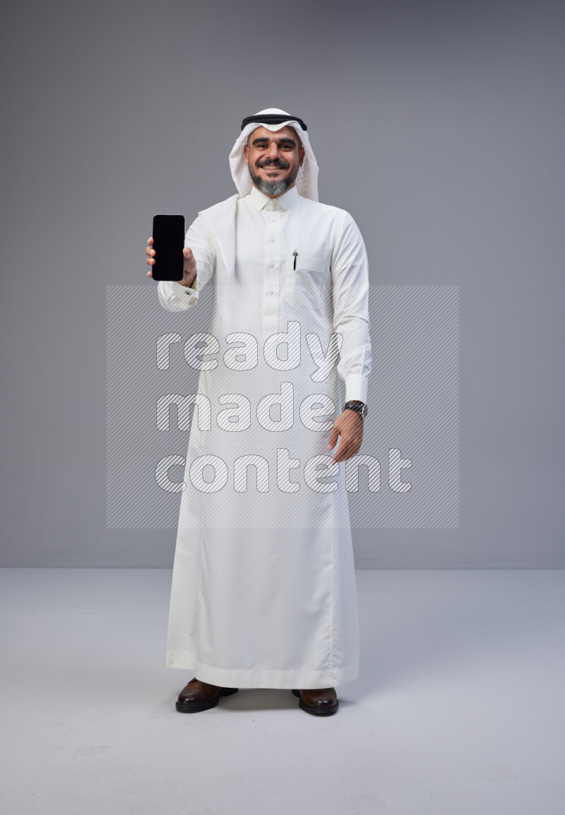 Saudi man Wearing Thob and white Shomag standing showing phone to camera on Gray background