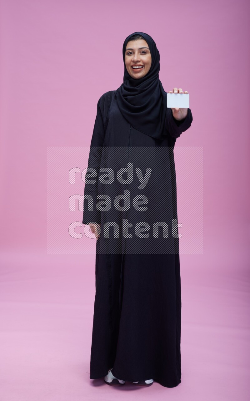 Saudi woman wearing Abaya standing holding ATM card on pink background