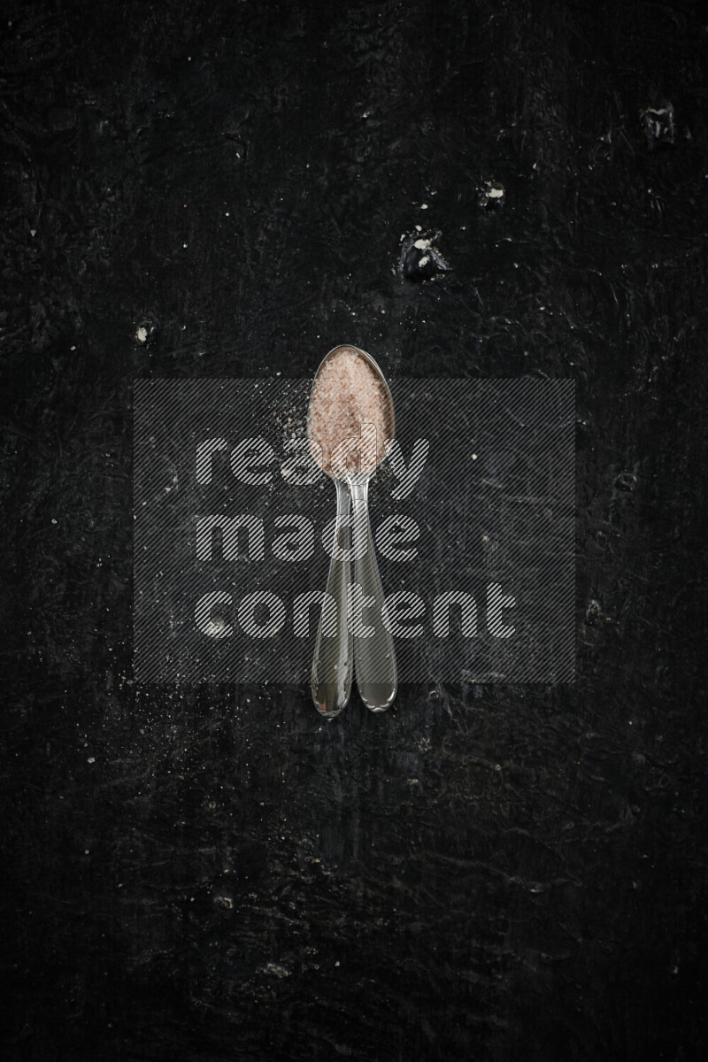 2 metal spoons filled with pink himalayan salt on black background