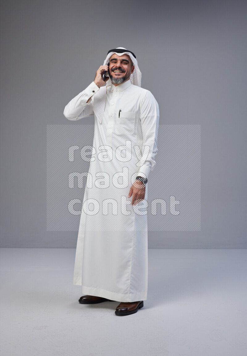 Saudi man Wearing Thob and white Shomag standing talking on phone on Gray background