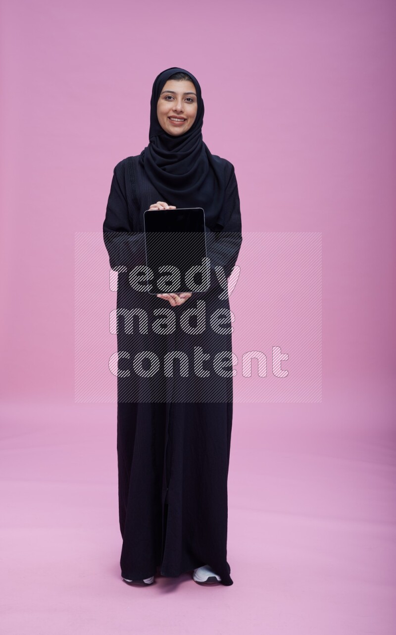 Saudi woman wearing Abaya standing showing tablet to camera on pink background