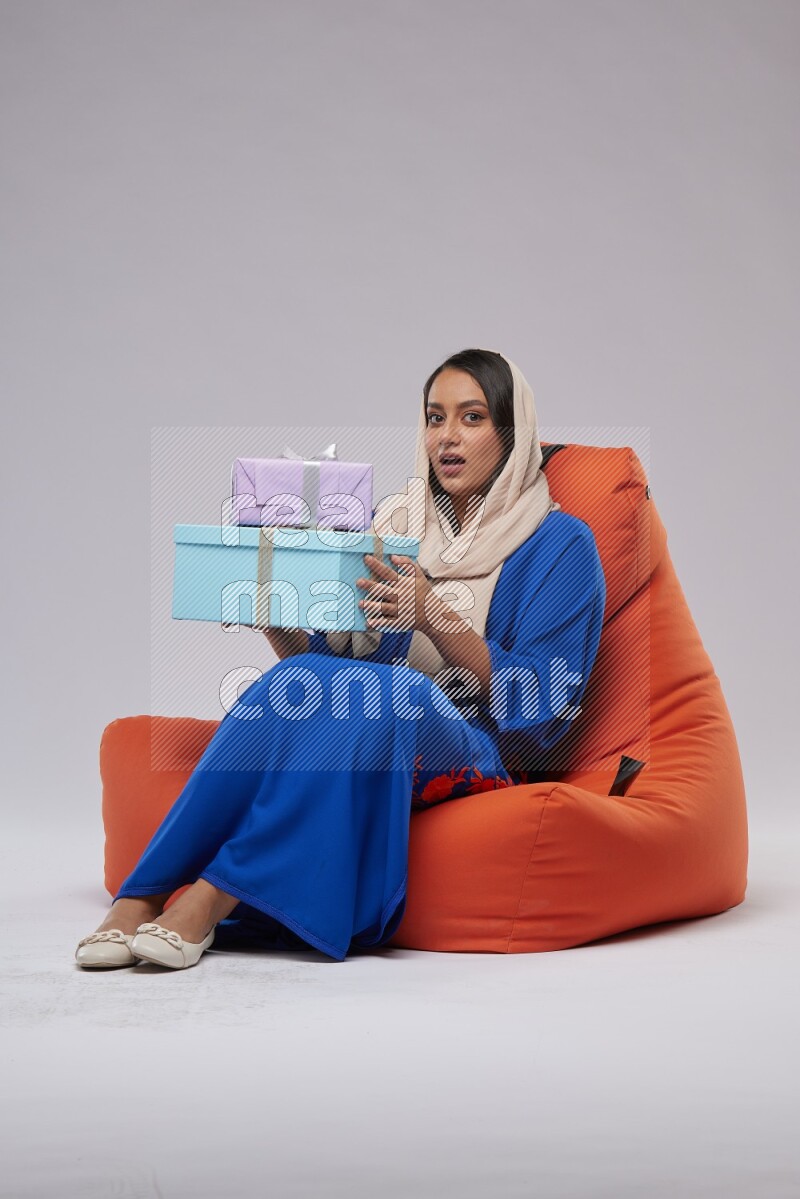 A woman sitting on an orange beanbag wearing Jalabeya holding a gift box