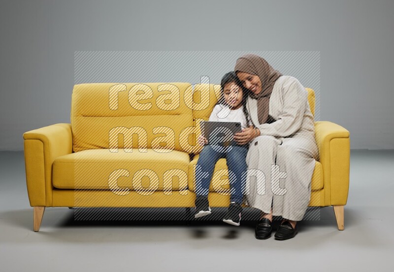 A girl with her mother on yellow sofa and watching on iPad on gray background